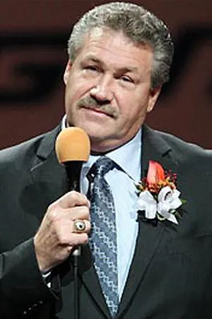 NHL Player Dave Shultz in a black suit with a orange lapel and holding a microphone.