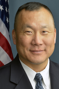 FBI Crisis Management expert Jin Kim in a suit and tie standing in front of an American flag.