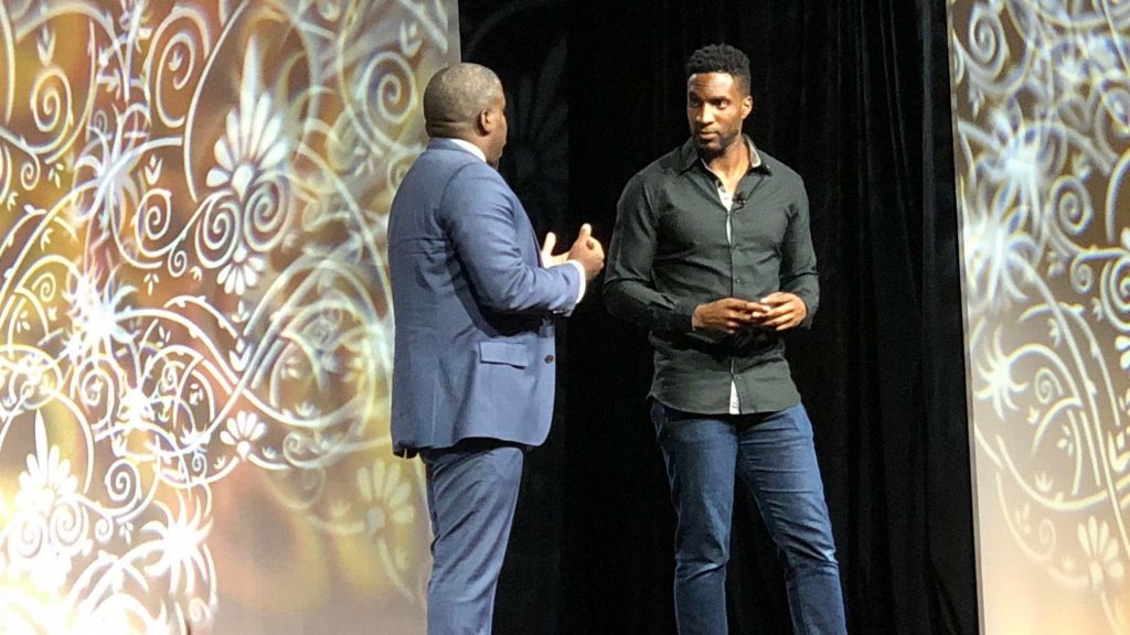 AK Ikwuakor in a black shirt standing on stage with another person in a suit.