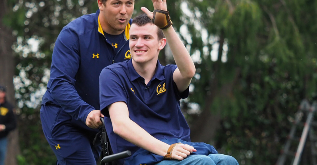 Rob Paylor being pushed in his wheelchair, smiling and waving.