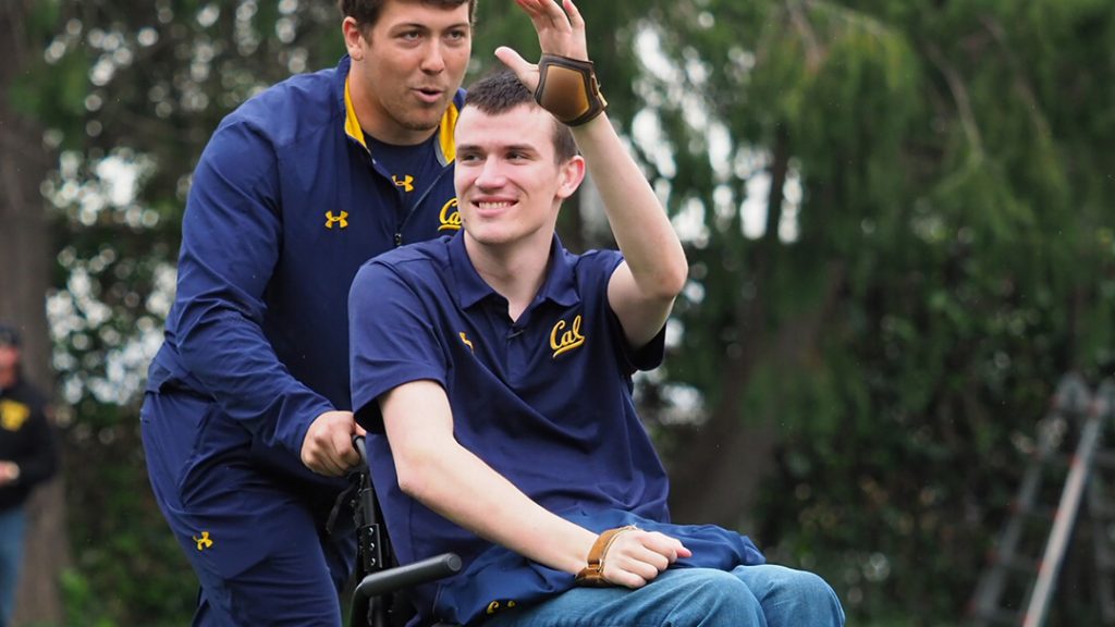 Two men, Robert Paylor in his wheelchair wearing his cal rugby shirt, waiving with his left hand. 