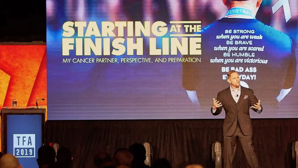 Matthew S. Newman speaking with a projection behind him fo the words "starting at the finish line"