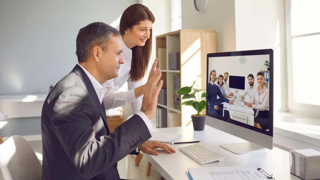 A man and a woman in business clothing waving at a computer displaying others on a video call for a virtual meeting
