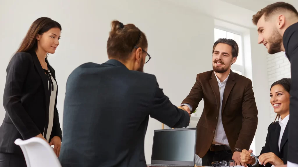 2 Men Shaking hands surounded by 3 others, all in business attire.