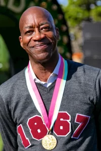 Olympian Edwin Moses smiling, wearing a medal and a shirt that says 1867