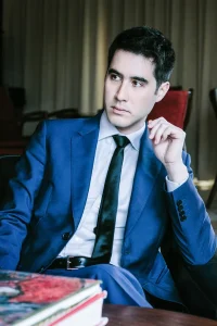 Magician and keynote speaker David Kwong in a blue suit, black tie, sitting down.